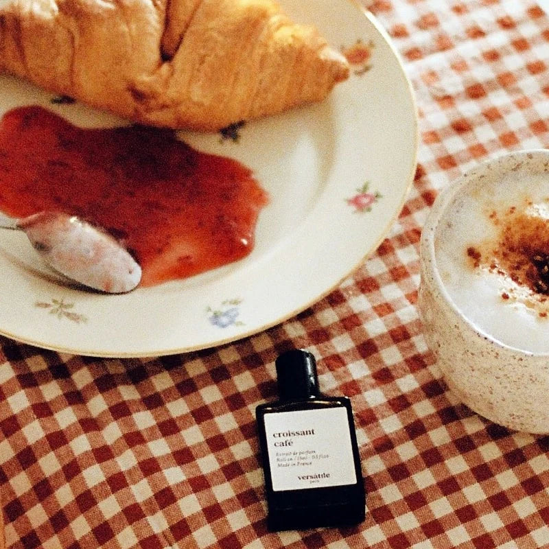 Bottle of croissant cafe perfume on a checkered tablecloth with a croissant and coffee.