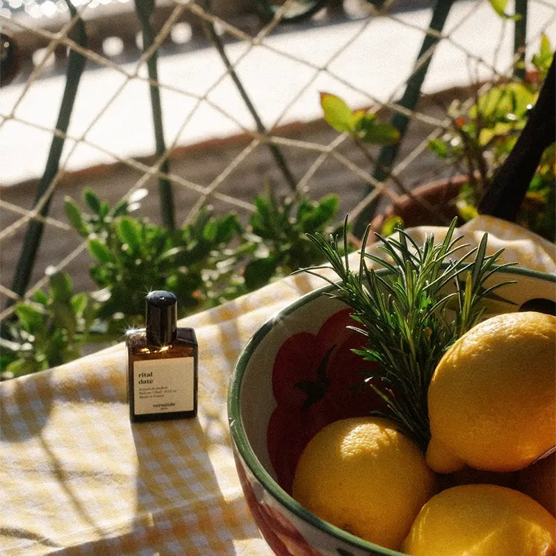 Bottle of olive oil next to a bowl of lemons and rosemary on a checkered tablecloth with plants in the background.