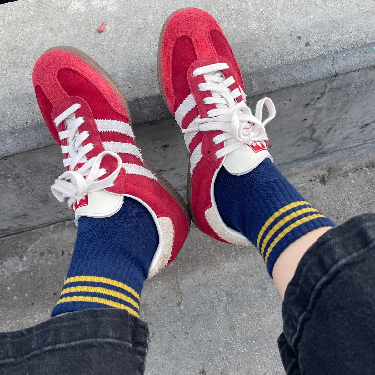 Red sneakers with white laces and blue socks with yellow stripes on a concrete surface.