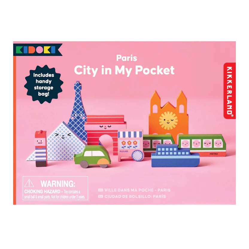 Paris City In My Pocket wooden toy set packaging. The box features a pink background with an illustrated display of the nine wooden pieces, including the Eiffel Tower, Notre Dame, a metro train, a car, a boat, and a flower shop. The text highlights the inclusion of a handy storage bag. A warning label indicates small parts, not suitable for children under 3 years.