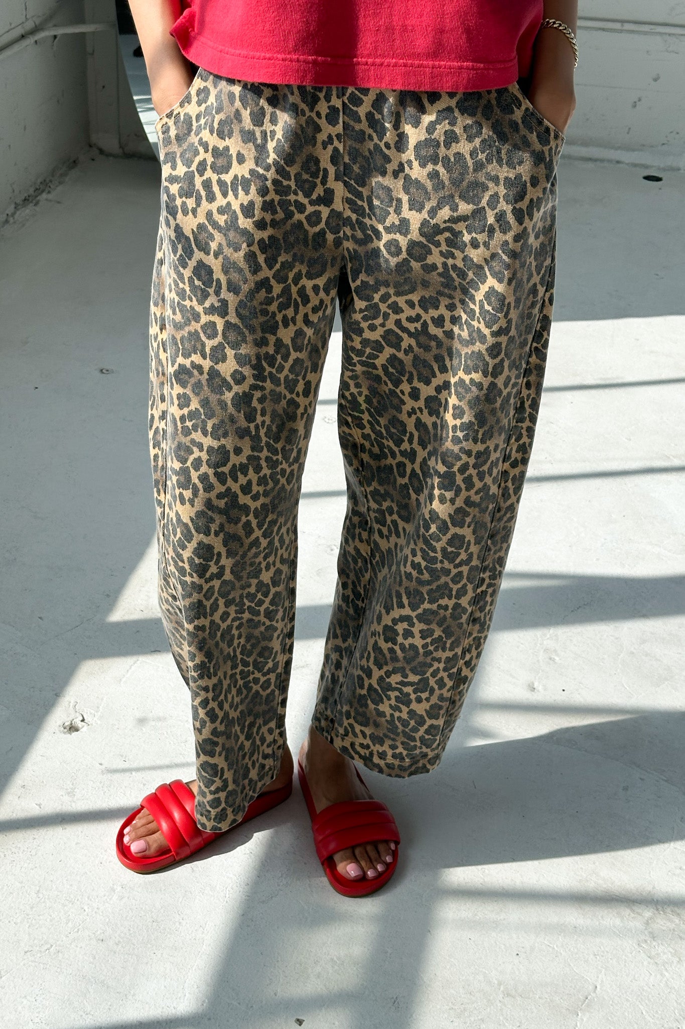 Close-up of model wearing Le Bon Shoppe Leopard Arc Pants with hands in pockets, paired with a red top and bright red slide sandals, standing in soft natural light on a concrete floor.