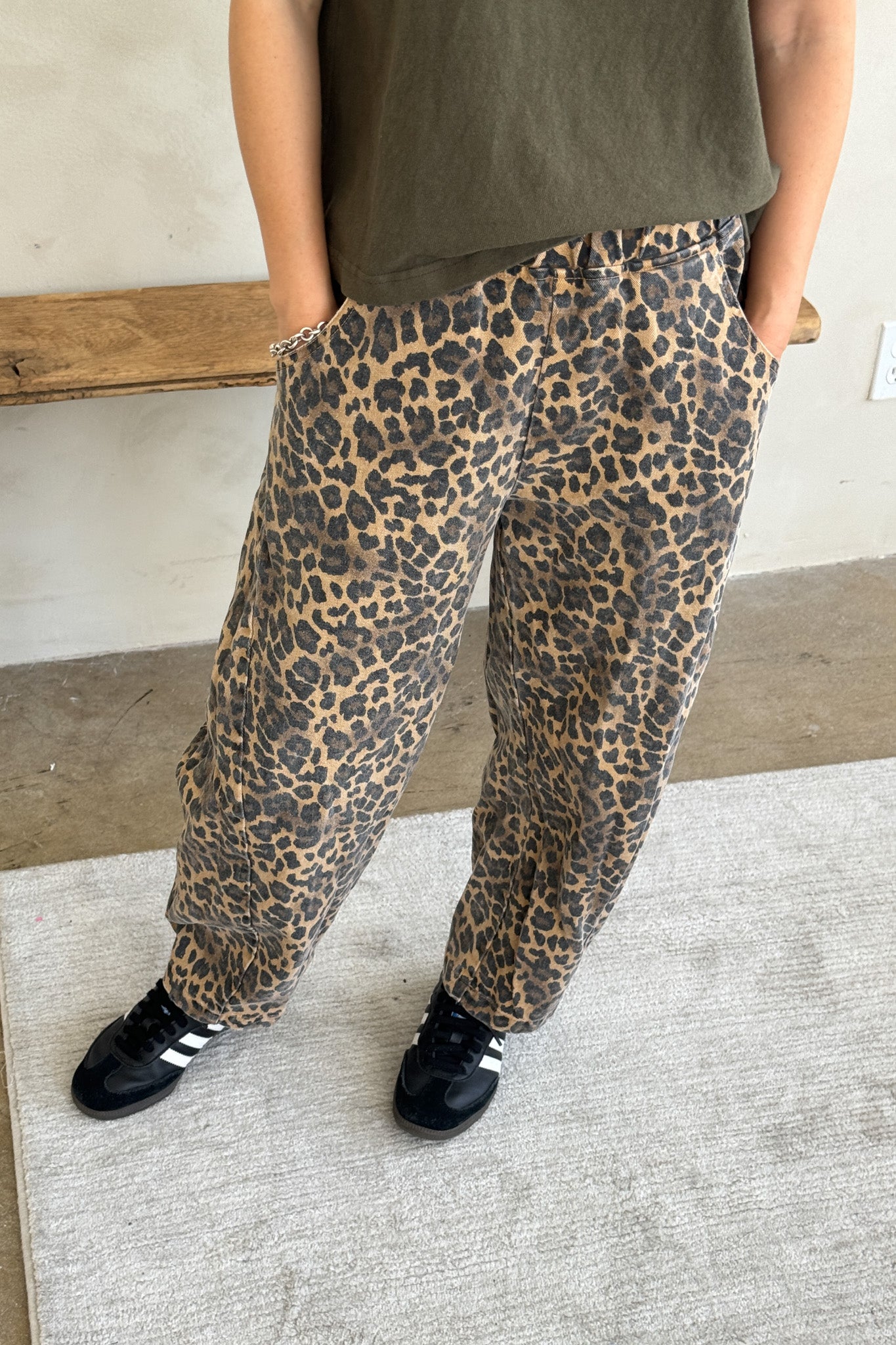 Close-up of model wearing Le Bon Shoppe Leopard Arc Pants with hands in pockets, paired with a khaki green T-shirt and black Adidas trainers, standing on a neutral rug with a wooden bench in the background.