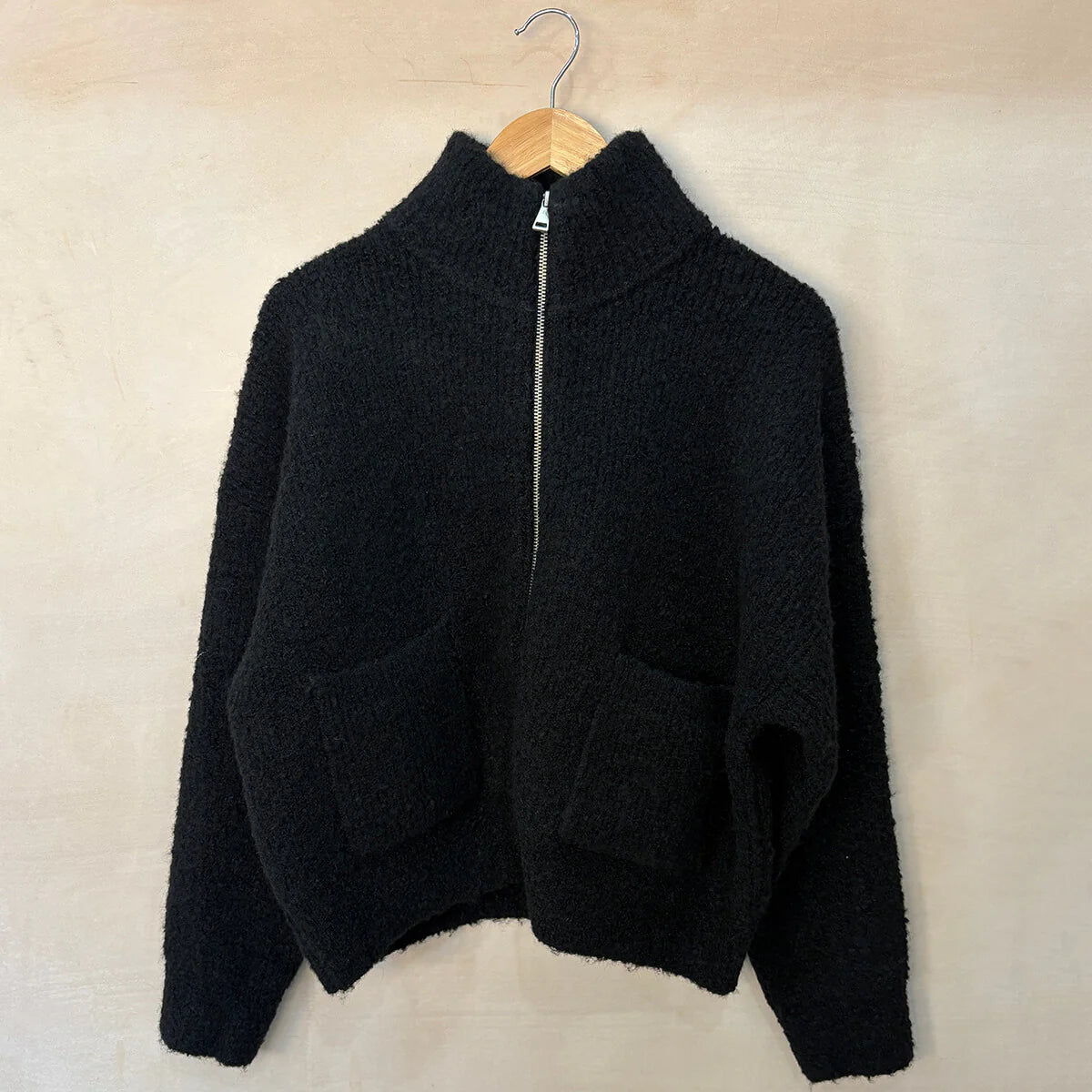Black zip-up sweater on a wooden hanger against a beige wall.