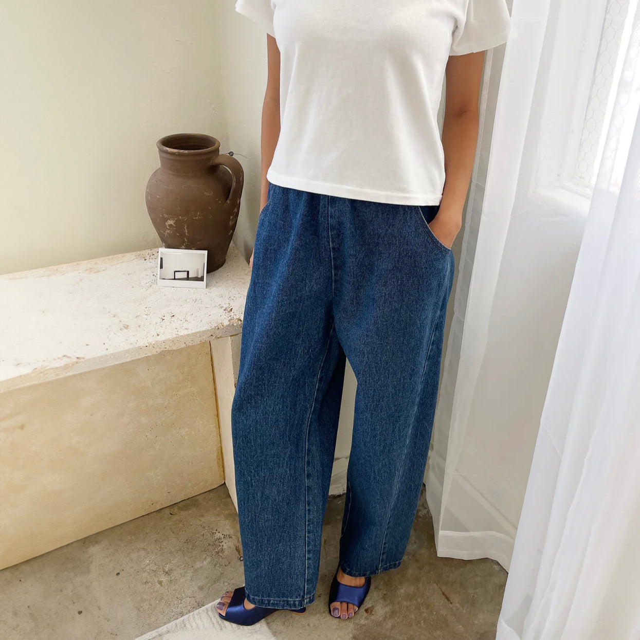 A pair of stylish high-waisted Arc Pants made from heavyweight denim, featuring a cropped length and slightly curved leg shape. The design includes unique side panels and deep pockets, perfect for casual wear. They are worn with a white t-shirt and blue sandals
