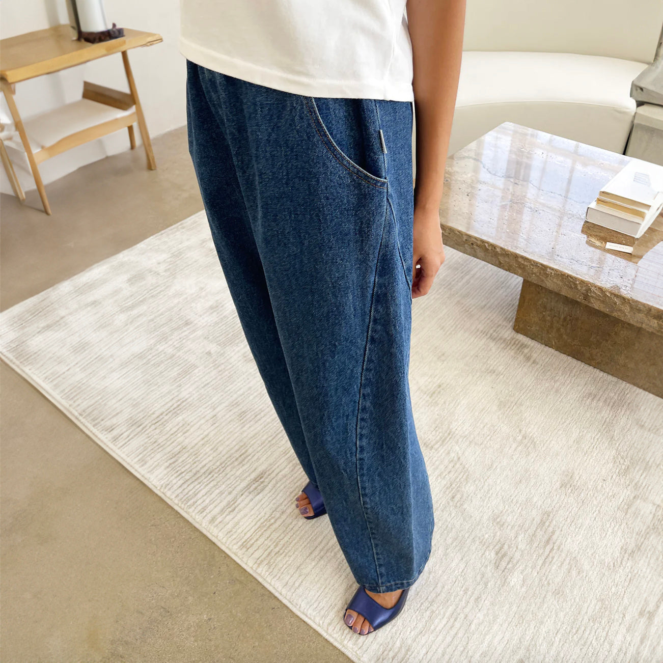 A pair of stylish high-waisted Arc Pants made from heavyweight denim, featuring a cropped length and slightly curved leg shape. The design includes unique side panels and deep pockets, perfect for casual wear. A side view looking don at the pants where the model wear blue sandals against a cream rug.