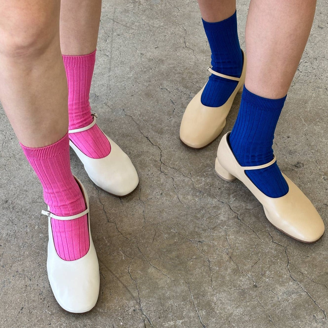 Two people wearing ribbed crew socks—one in cobalt blue and the other in bright pink—paired with beige and white Mary Jane heels, standing on a concrete floor.