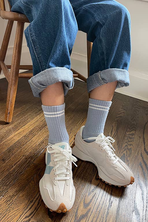 A person wearing light blue ribbed socks with white stripes at the cuffs, paired with rolled-up denim jeans and white sneakers, sitting on a wooden floor.