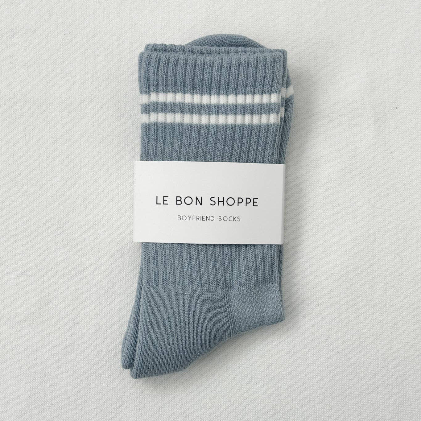 A pair of ribbed grey socks featuring red and blue stripes at the cuffs and "Le Bon Shoppe" branding on the foot, laid flat on a textured surface.