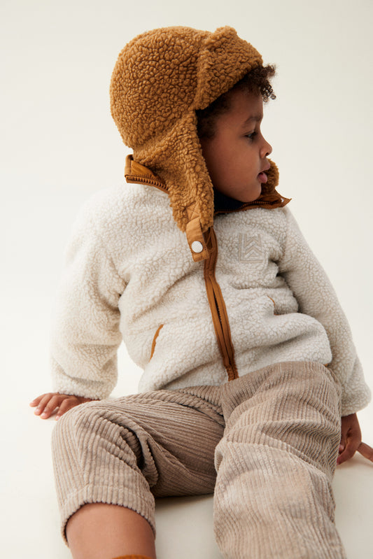 Liewood Golden Caramel Bravo Pile Hat for children, featuring soft fleece lining, protective earflaps, and adjustable snap-button straps. The model looks to the side in a cream fleece jacket and beige cords