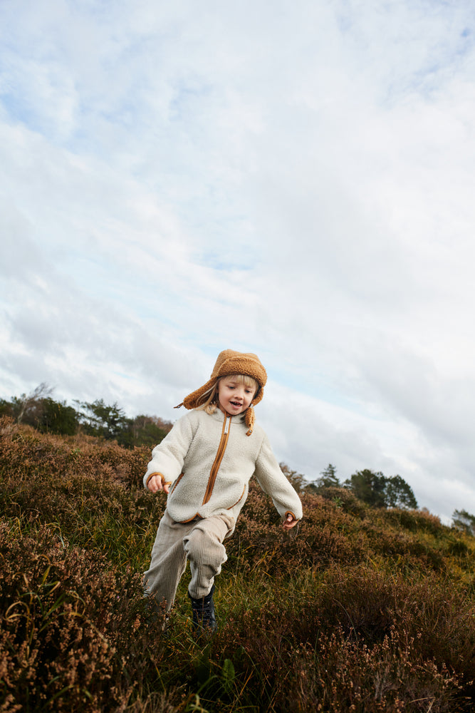 Liewood Golden Caramel Bravo Pile Hat for children, featuring soft fleece lining, protective earflaps, and adjustable snap-button straps. The model runs outside in a cream fleece jacket and beige cords