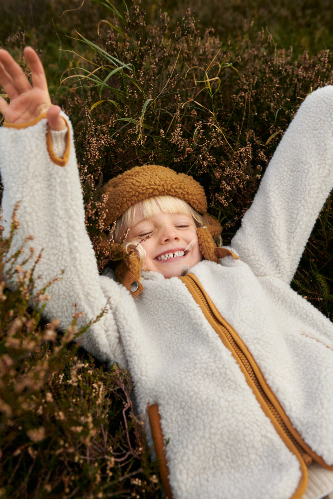 Liewood Golden Caramel Bravo Pile Hat for children, featuring soft fleece lining, protective earflaps, and adjustable snap-button straps. The model lays on some heather outside in a cream fleece jacket