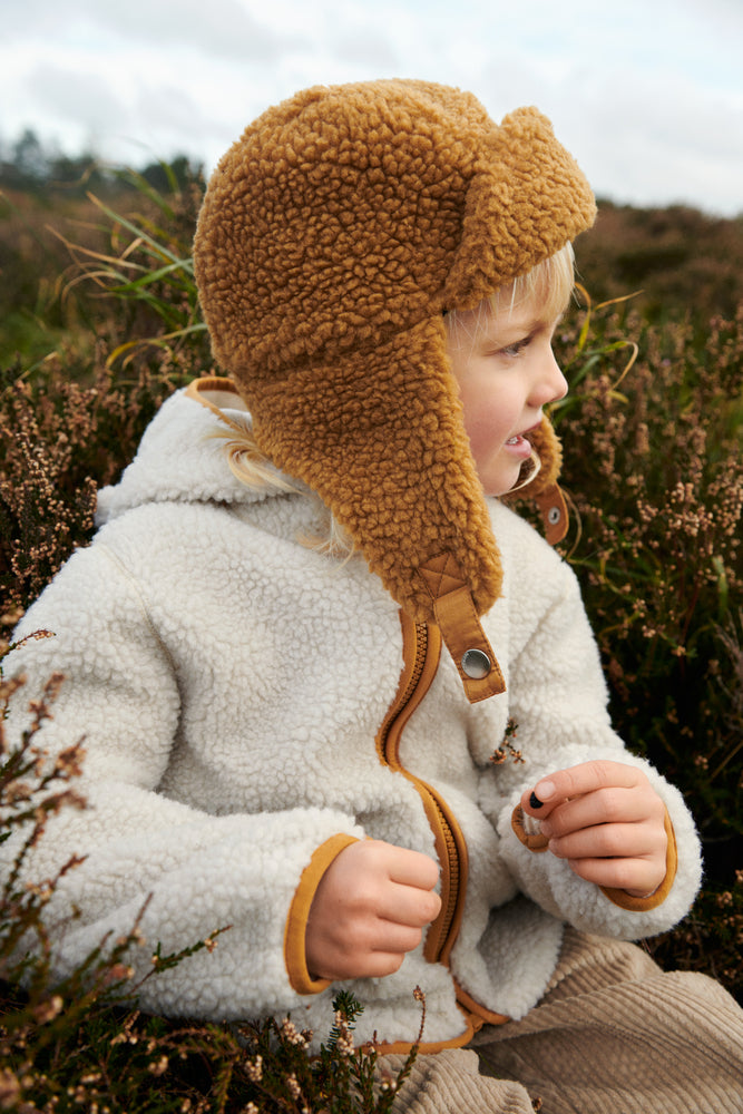 Liewood Golden Caramel Bravo Pile Hat for children, featuring soft fleece lining, protective earflaps, and adjustable snap-button straps. The model looks to the side in a cream fleece jacket and beige cords