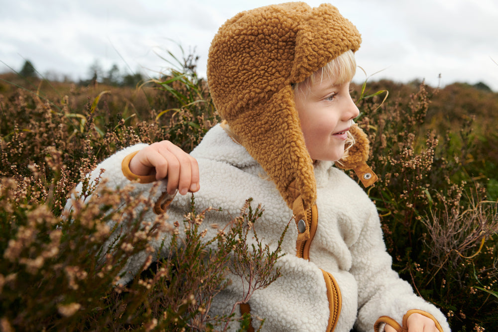 Liewood Golden Caramel Bravo Pile Hat for children, featuring soft fleece lining, protective earflaps, and adjustable snap-button straps. The model looks to the side in a cream fleece jacket