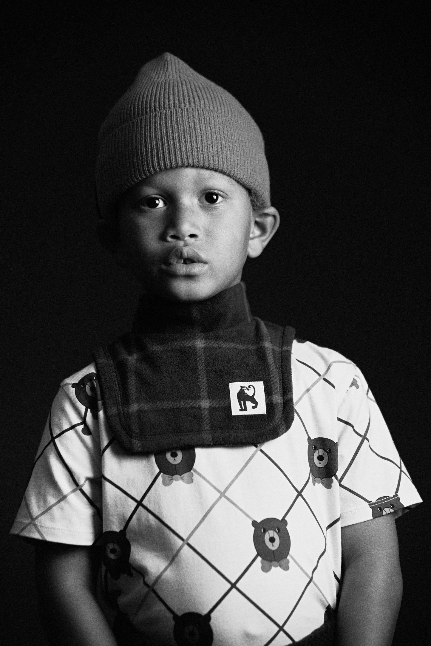 Black and white photo of a child wearing a patterned shirt and knitted hat.