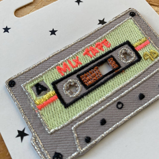 A detailed close-up of the patch, showcasing intricate metallic silver stitching, bold black cassette reels, and vibrant orange and yellow stripe accents, giving it an authentic vintage mixtape aesthetic.