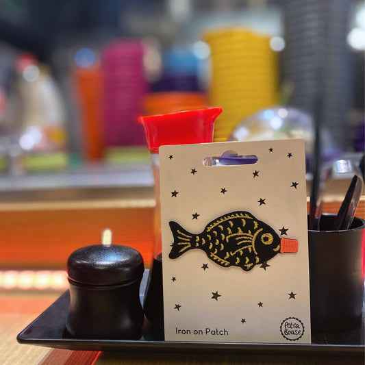 The embroidered patch displayed on its packaging, propped up on a restaurant counter surrounded by chopsticks, condiments, and blurred colourful tableware, adding a playful nod to its sushi bar inspiration.