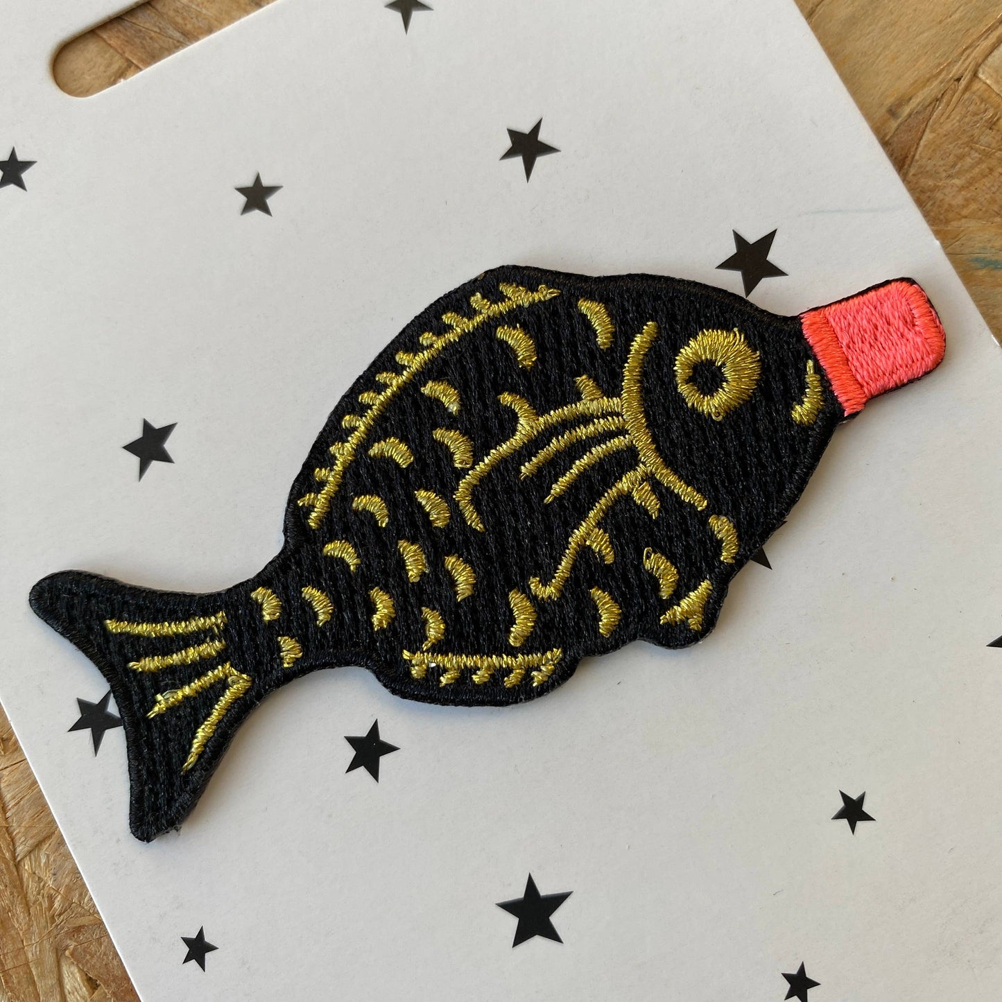 A detailed close-up of the embroidered patch, showcasing the gold stitched fish scales, textured black body, and vibrant red bottle cap against a white background with scattered black stars.