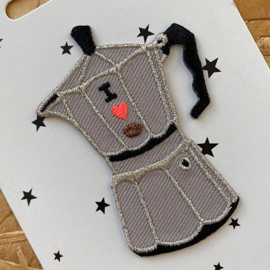 A detailed close-up of the embroidered patch, highlighting the moka pot's structured silver panels, bold black handle, and intricate embroidered details, including an "I ❤️ ☕" design stitched onto the front.