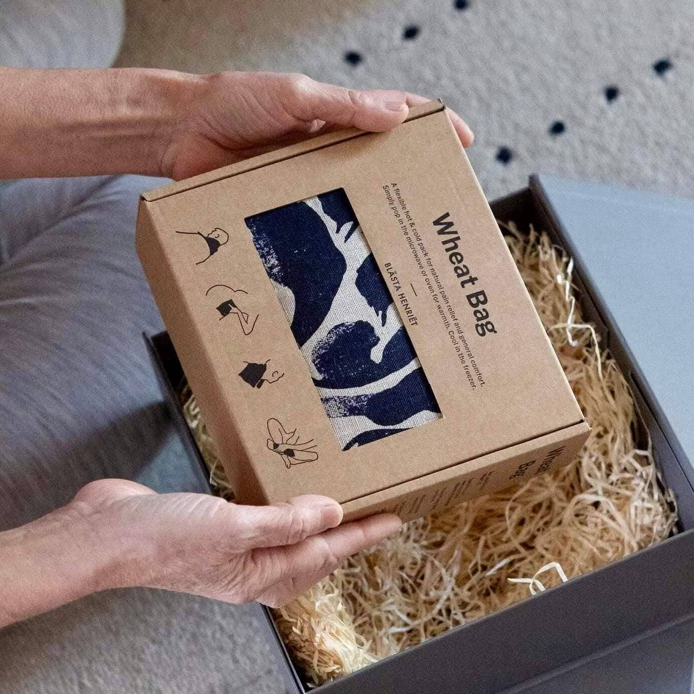 Person holding a cardboard box with a wheat bag product inside, set against a neutral background.