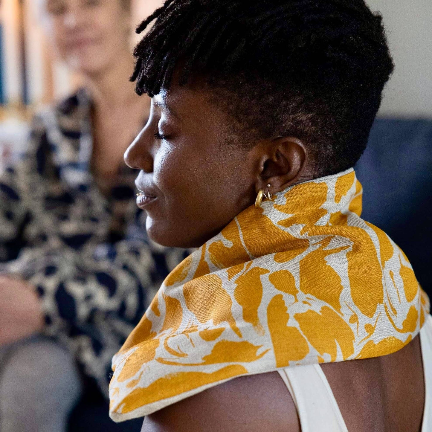Woman wearing a yellow and white patterned wheat bag, with another person partially visible in the background.