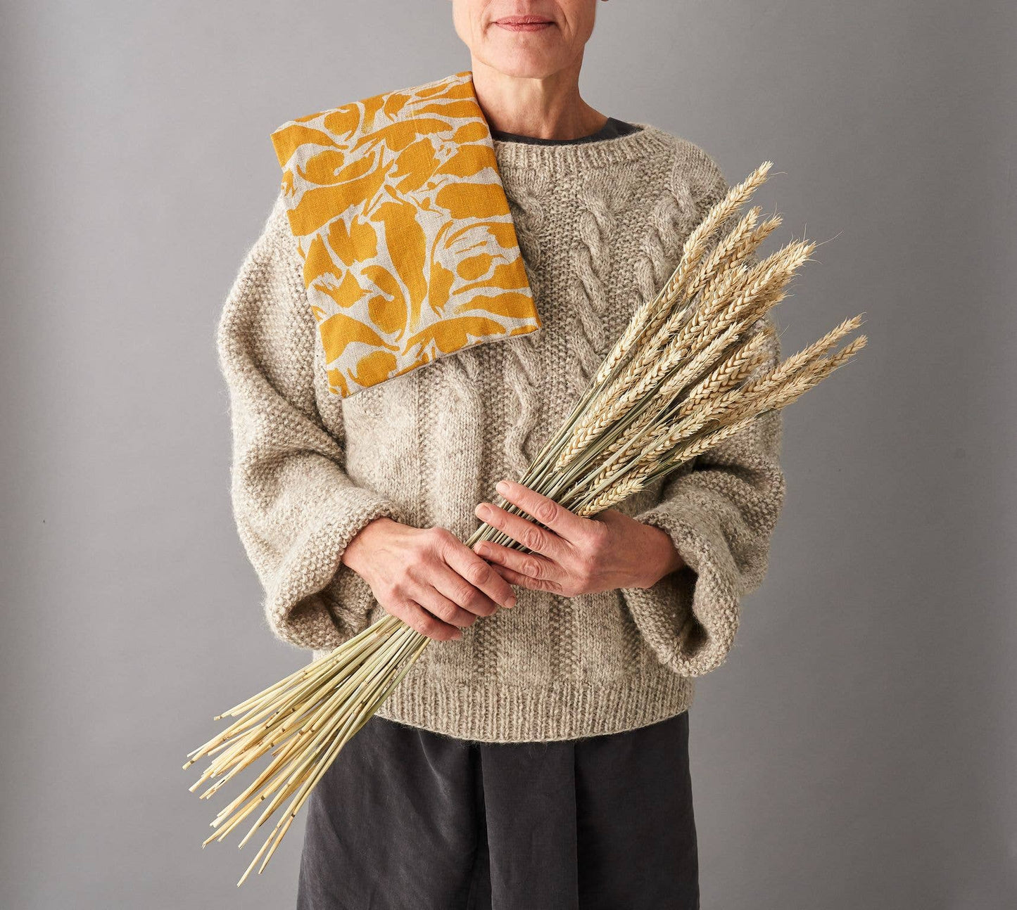 Person wearing a beige sweater holding a bunch of wheat with a yellow patterned wheat bag draped over their shoulder against a gray background.