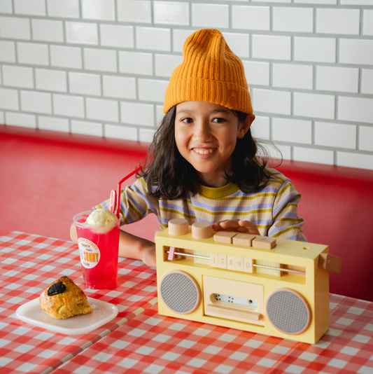 Child wearing a yellow beanie sitting at a table with a toy boombox, drink, and dessert.