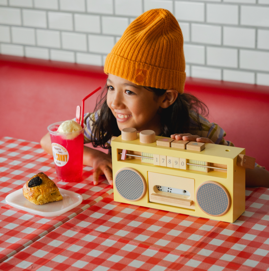 Child with a yellow beanie sitting at a table with a toy boombox, ice cream, and dessert.