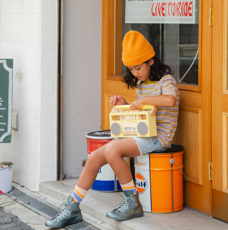Person sitting on a stack of oil drums with a yellow boombox, wearing a yellow beanie and striped shirt, in front of a store entrance.