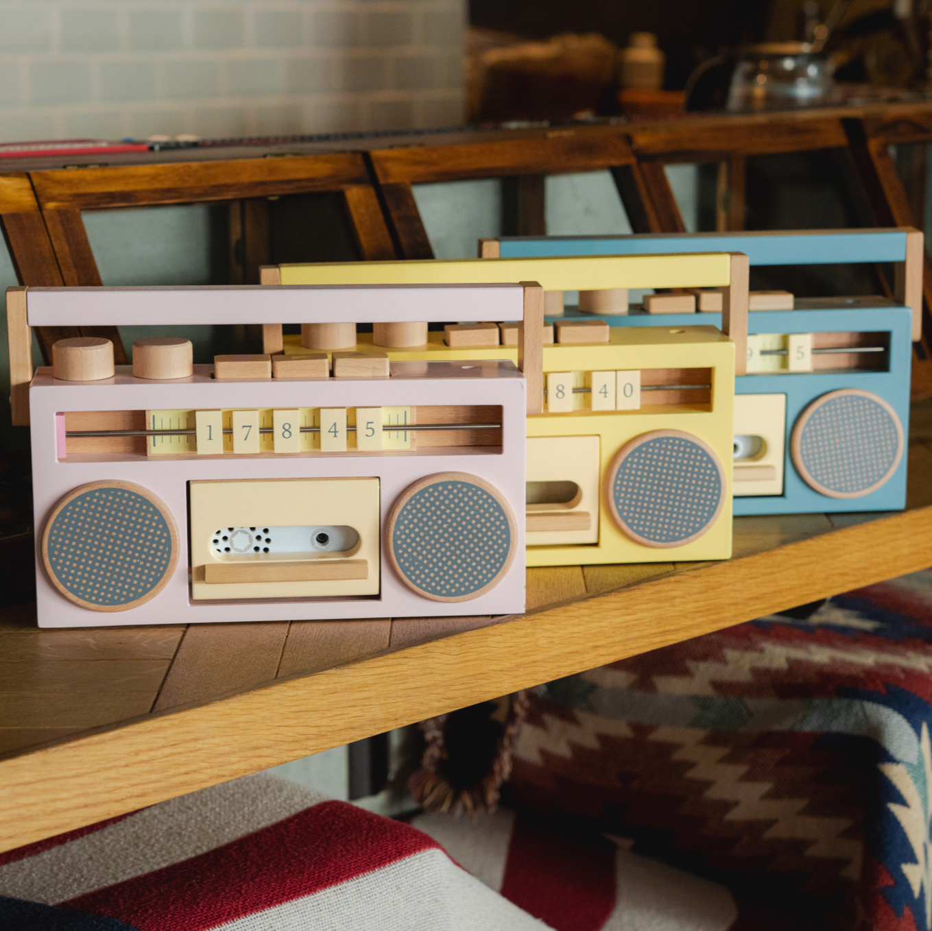 Colorful wooden boombox models on a wooden shelf with a patterned rug and furniture in the background.