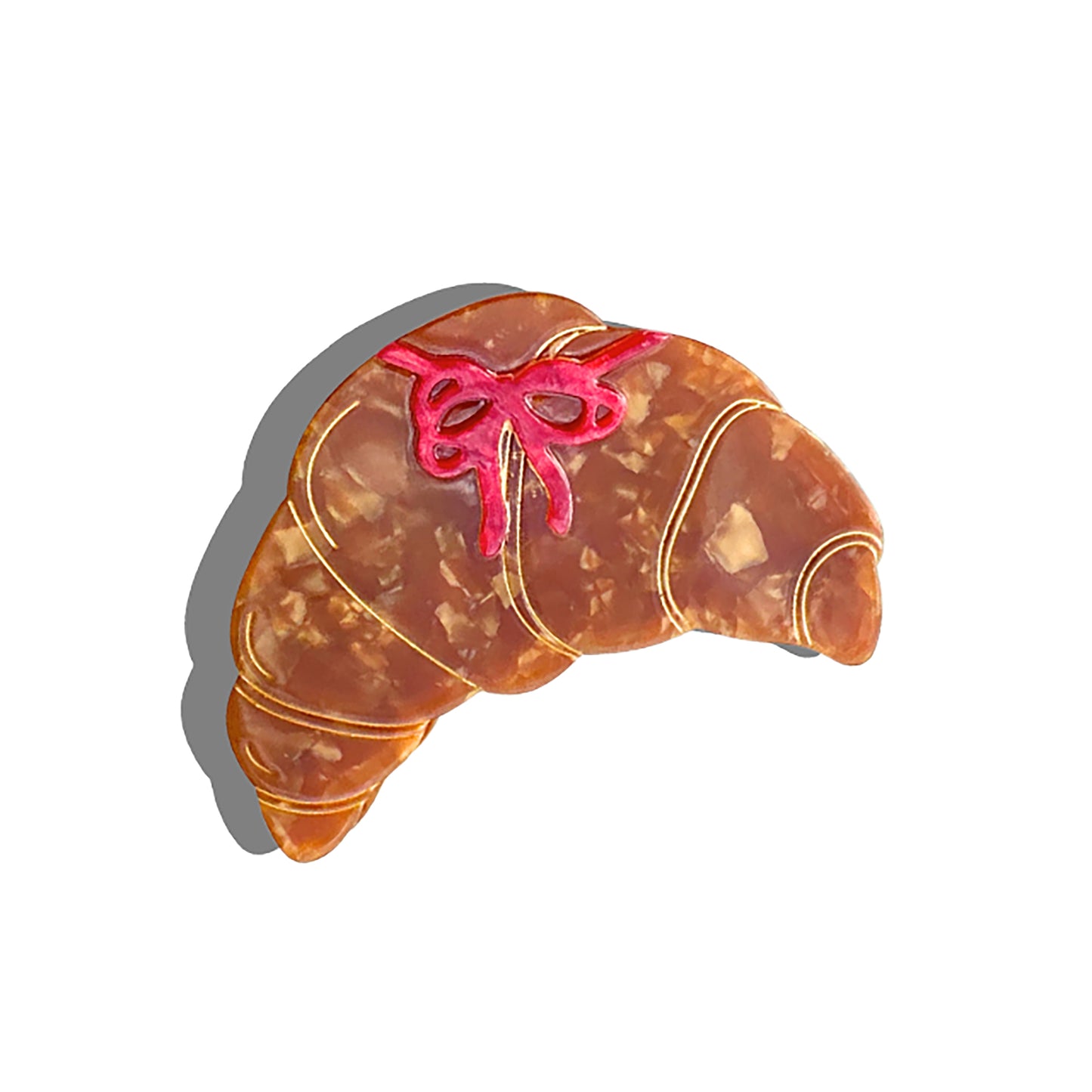 A croissant-shaped hair claw with a marbled brown and beige pattern, featuring delicate gold detailing that outlines the pastry layers. A small red bow design adds a playful touch. The accessory casts a soft shadow, enhancing its three-dimensional appearance.