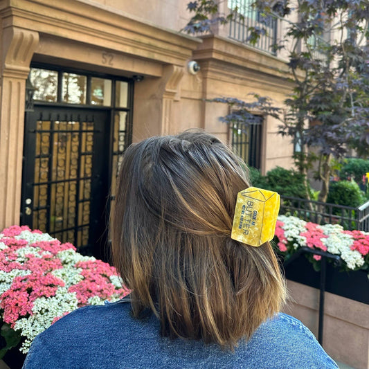 A person with shoulder-length brown hair wears a butter-shaped hair claw, securing part of their hair in a half-up style. The claw’s bright yellow marbled texture and 'Grade AA Butter' text stand out against the person's dark denim jacket. The background features a brownstone building with black iron gates, green window planters, and pink and white flowers.