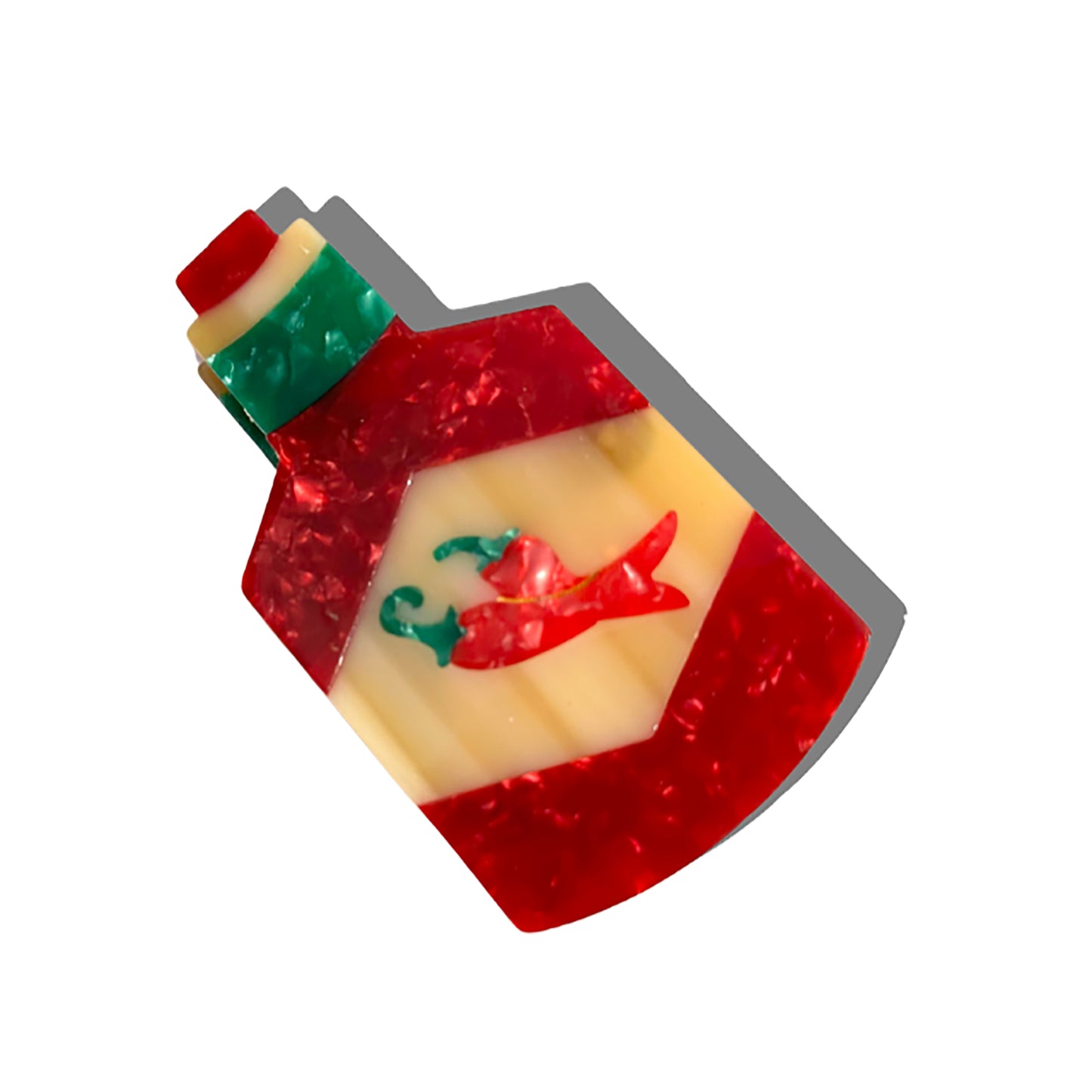 A close-up of the Solar Eclipse hand-painted hot sauce bottle hair claw clip, shaped like a bottle with a red, cream, and green design featuring embossed chili peppers. Made from eco-friendly cellulose acetate, it casts a soft shadow on a white background.