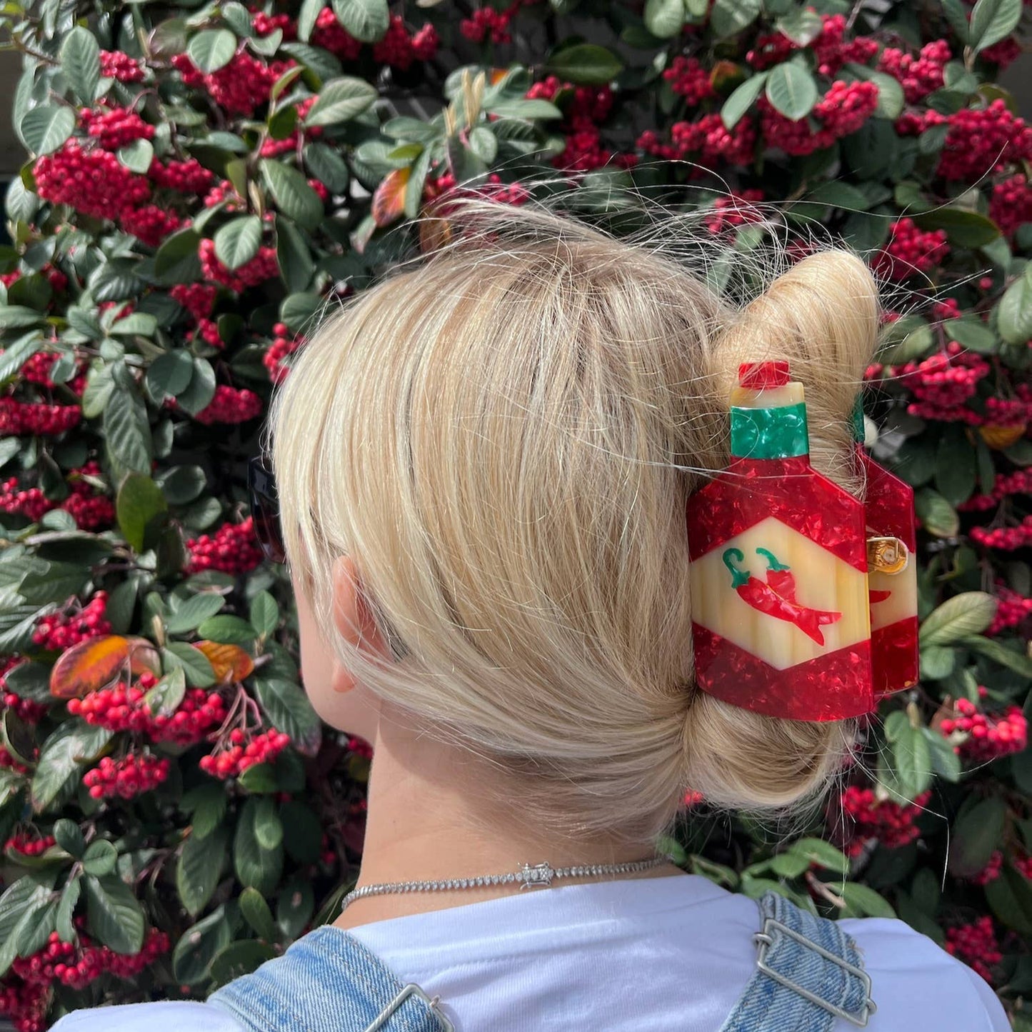A person wearing the hand-painted hot sauce bottle hair claw clip, securing their blonde hair in a twisted bun. The bold red and cream clip features a chili pepper design and is set against a lush backdrop of green foliage and red berries.