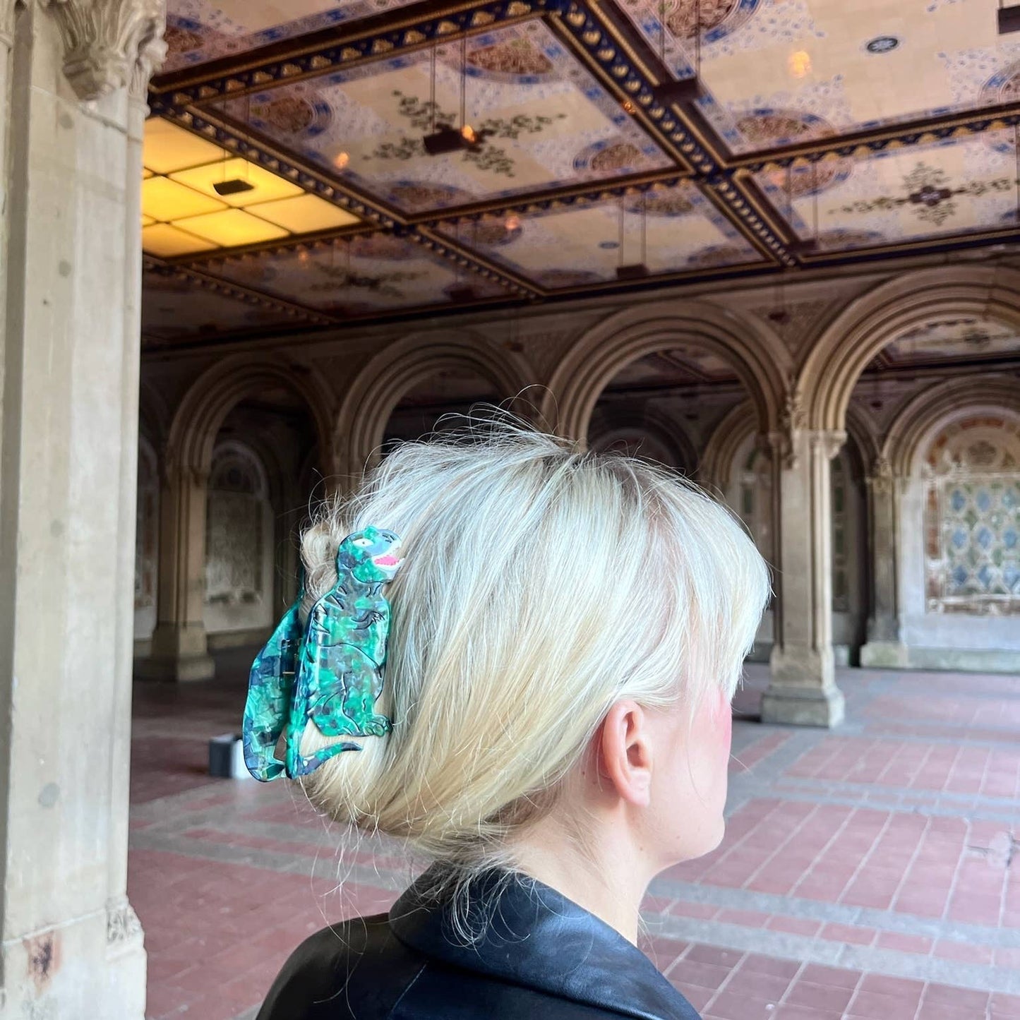 A person with blonde hair styled in a twisted bun, secured with the hand-painted T-Rex claw clip. They stand under an ornate, historic ceiling with intricate patterns and gold detailing, highlighting the clip’s unique and artistic design.