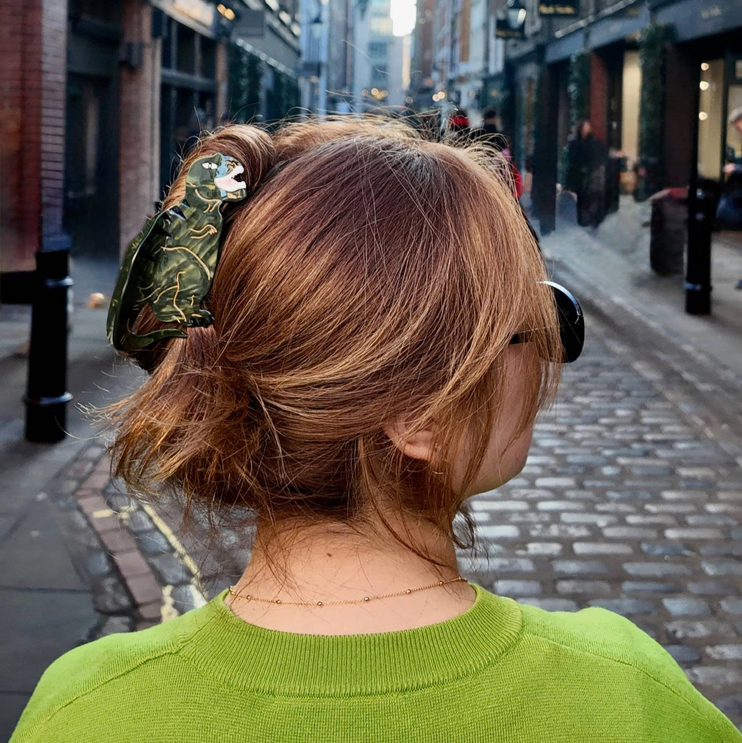A person wearing the T-Rex claw clip in a loose updo while walking down a cobbled street lined with brick buildings. The bold dinosaur design adds a playful touch to their look, standing out against their green sweater.