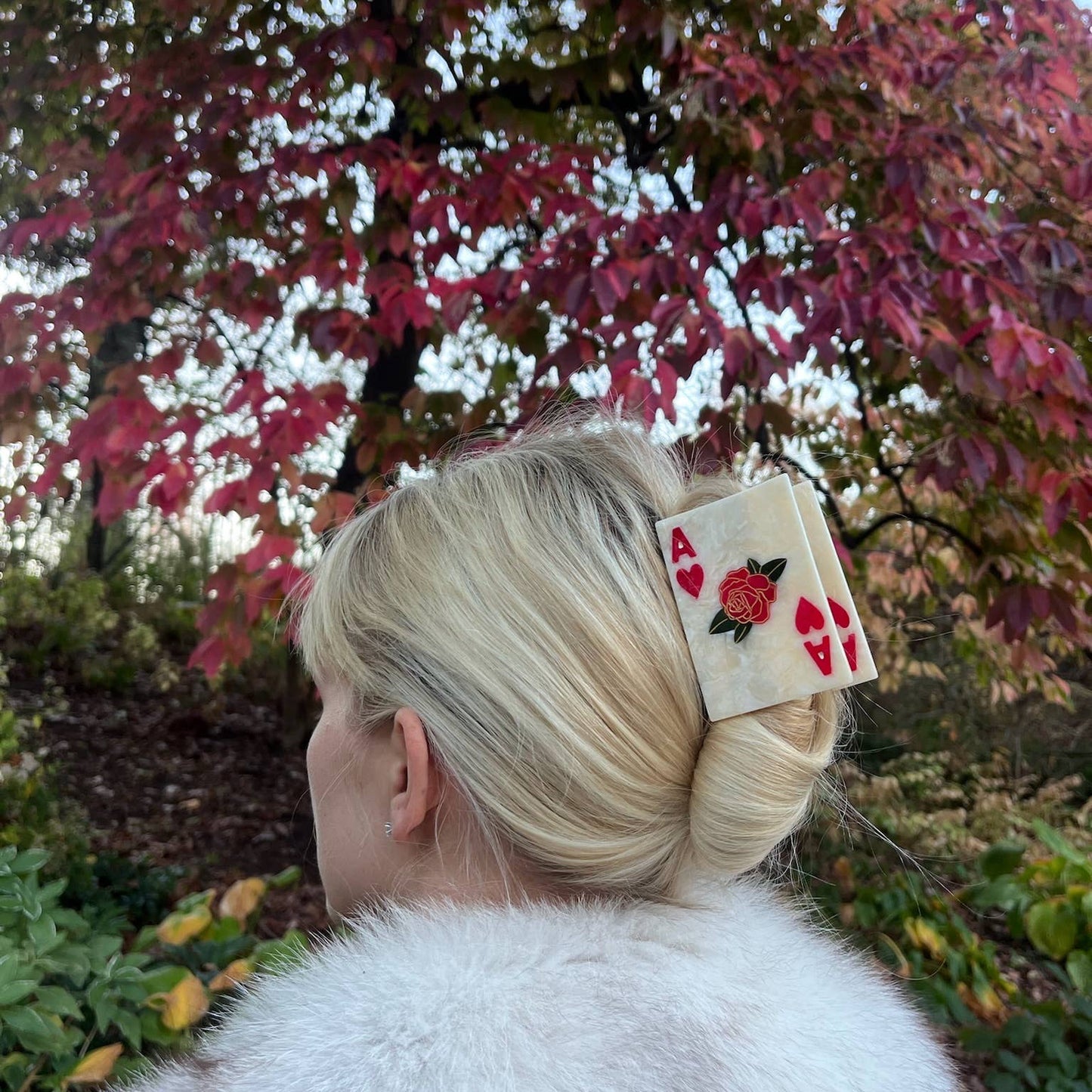 A woman with blonde hair styled in an elegant French twist wears an Ace of Hearts playing card-shaped hair claw, securing her updo with a bold and playful touch. The clip features a marbled ivory finish with red heart symbols and a detailed rose illustration in the centre. She is dressed in a luxurious white faux fur coat, standing against a backdrop of autumnal trees with red and green leaves.