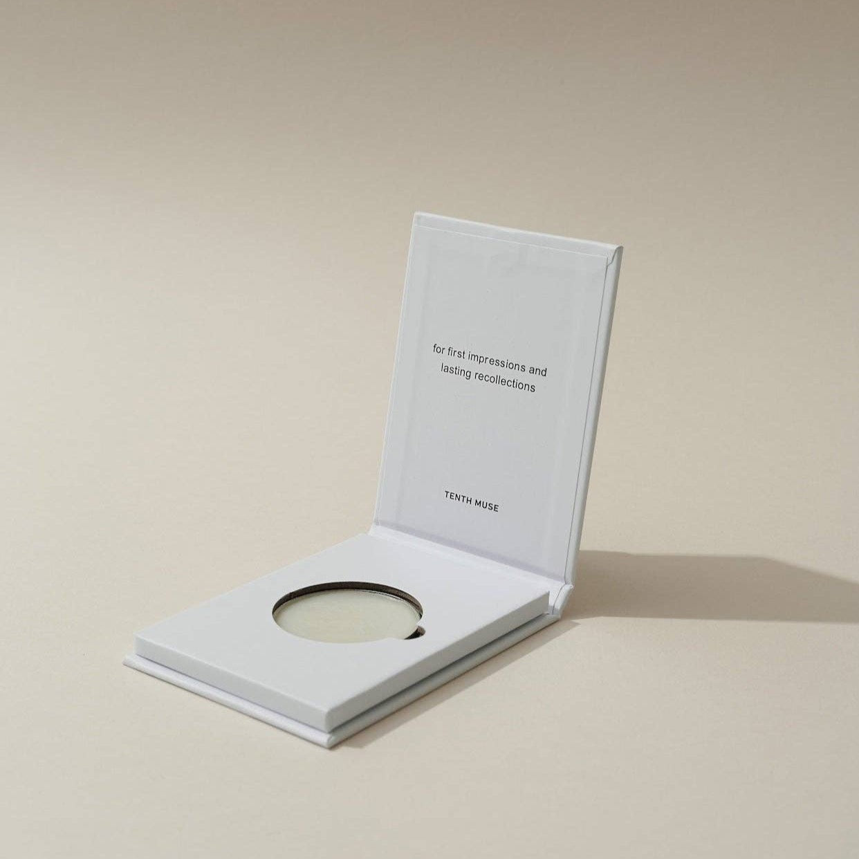Inside of the white sqaure packaging for the solid perfume balm. The inside lid says 'For first impressions and lasting recollections' in black box letters. Beneath that it says 'TENTH MUSE' also in black.