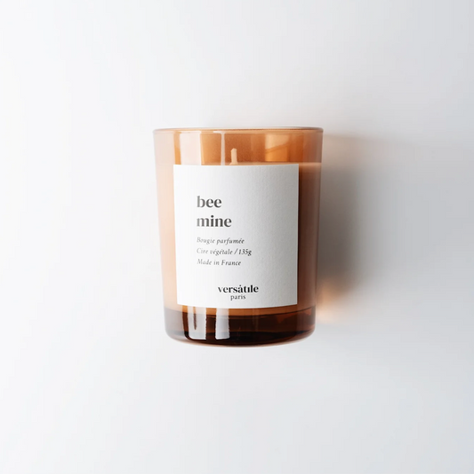 A beige candle with a label that reads 'Bee Mine' by Versatile Paris, with a clear glass jar and a white label, on a white background.