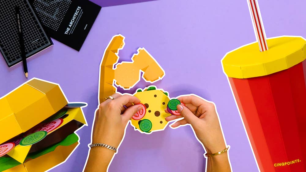 Hands assembling a cheeseburger model with a drink on a purple background