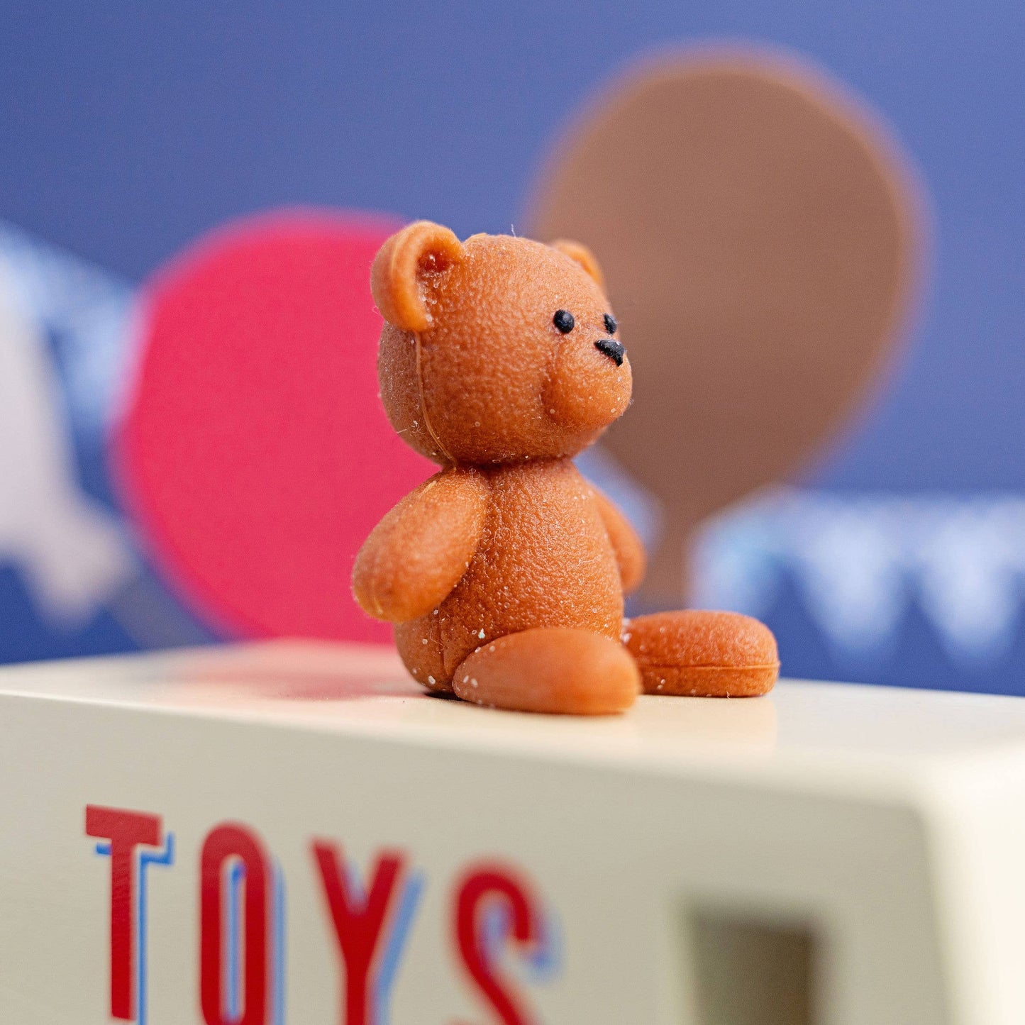 Small brown teddy bear figurine on a white surface with a colorful blurred background