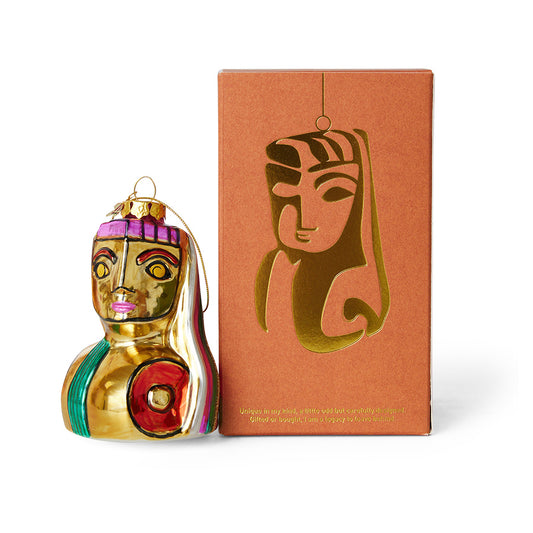 A golden Christmas ornament with a face and body, placed in front of its brown packaging box with a gold design on the top.