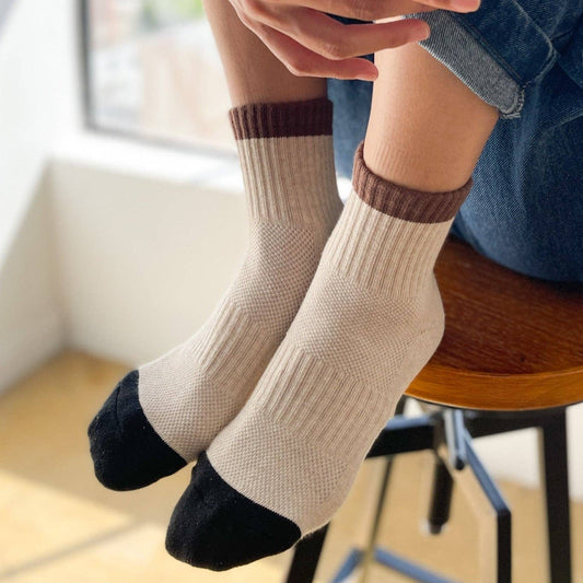 A person seated on a stool wearing a pair of Le Bon Shoppe Girlfriend Socks with color-blocked design, featuring a beige body with dark brown bands around the ankle and top, and a black toe cap.