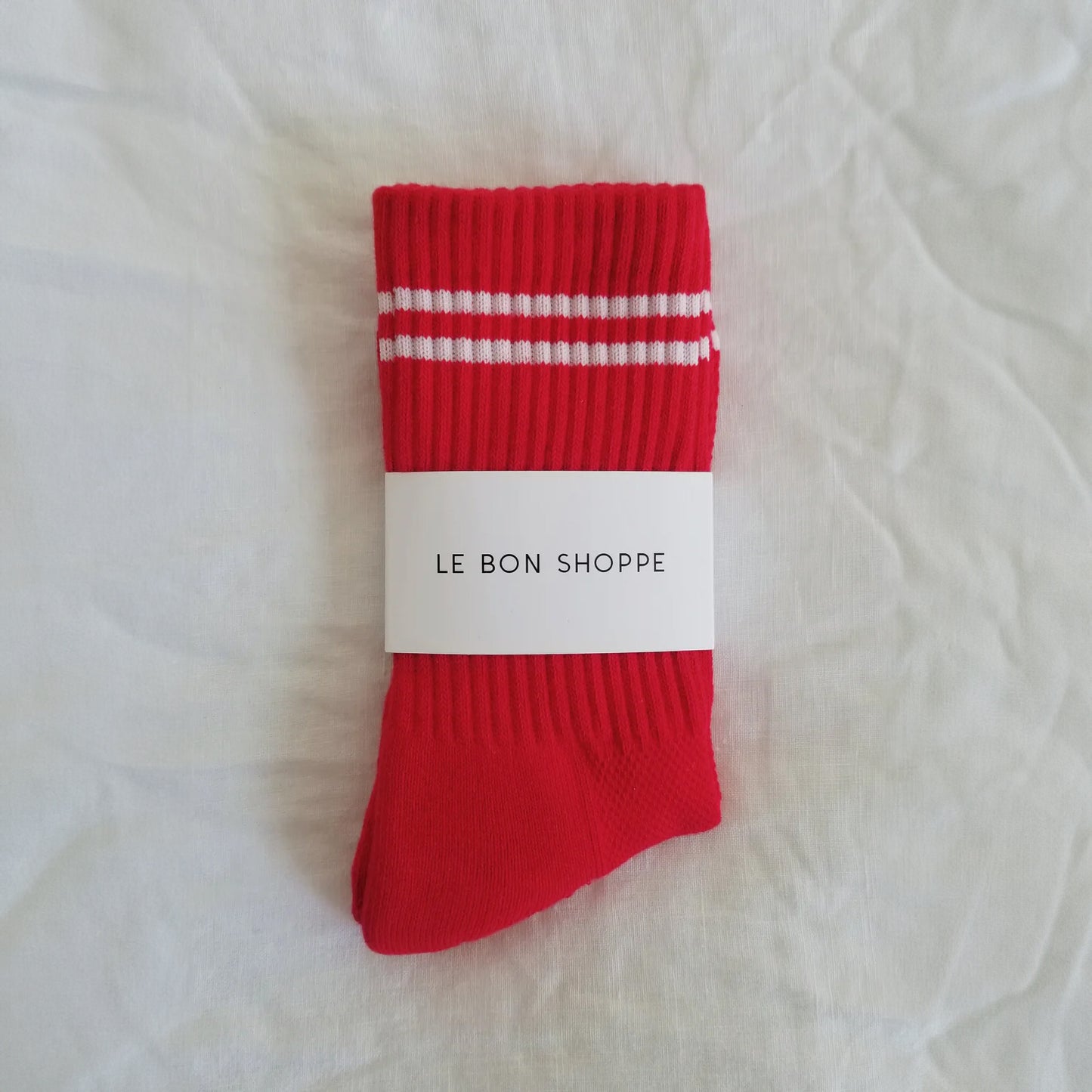 A pair of red ribbed crew socks with two white stripes near the cuff, neatly folded and wrapped with a white Le Bon Shoppe label, placed on a white fabric background.