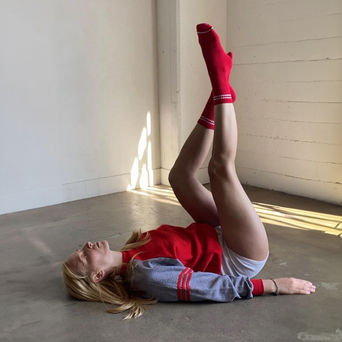 A woman lying on a concrete floor, stretching her legs up, wearing red ribbed crew socks with white stripes, paired with a red and grey sweatshirt.
