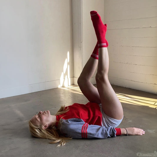 A woman lying on a concrete floor, stretching her legs up, wearing red ribbed crew socks with white stripes, paired with a red and grey sweatshirt.
