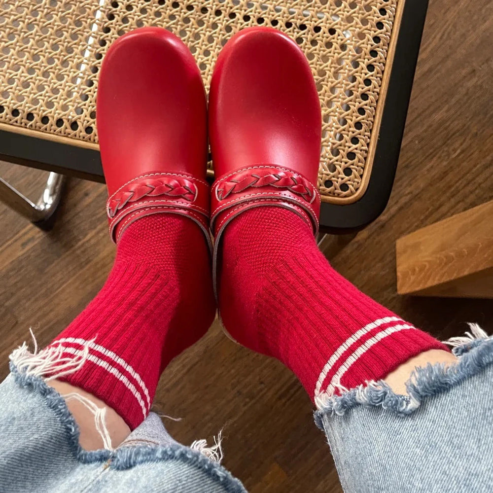 A person wearing red ribbed crew socks with white stripes, paired with matching red leather clogs, sitting on a woven chair with frayed denim jeans.