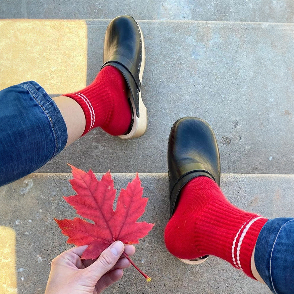A person wearing red ribbed crew socks with white stripes, paired with black leather clogs, sitting on outdoor steps while holding a red autumn leaf.