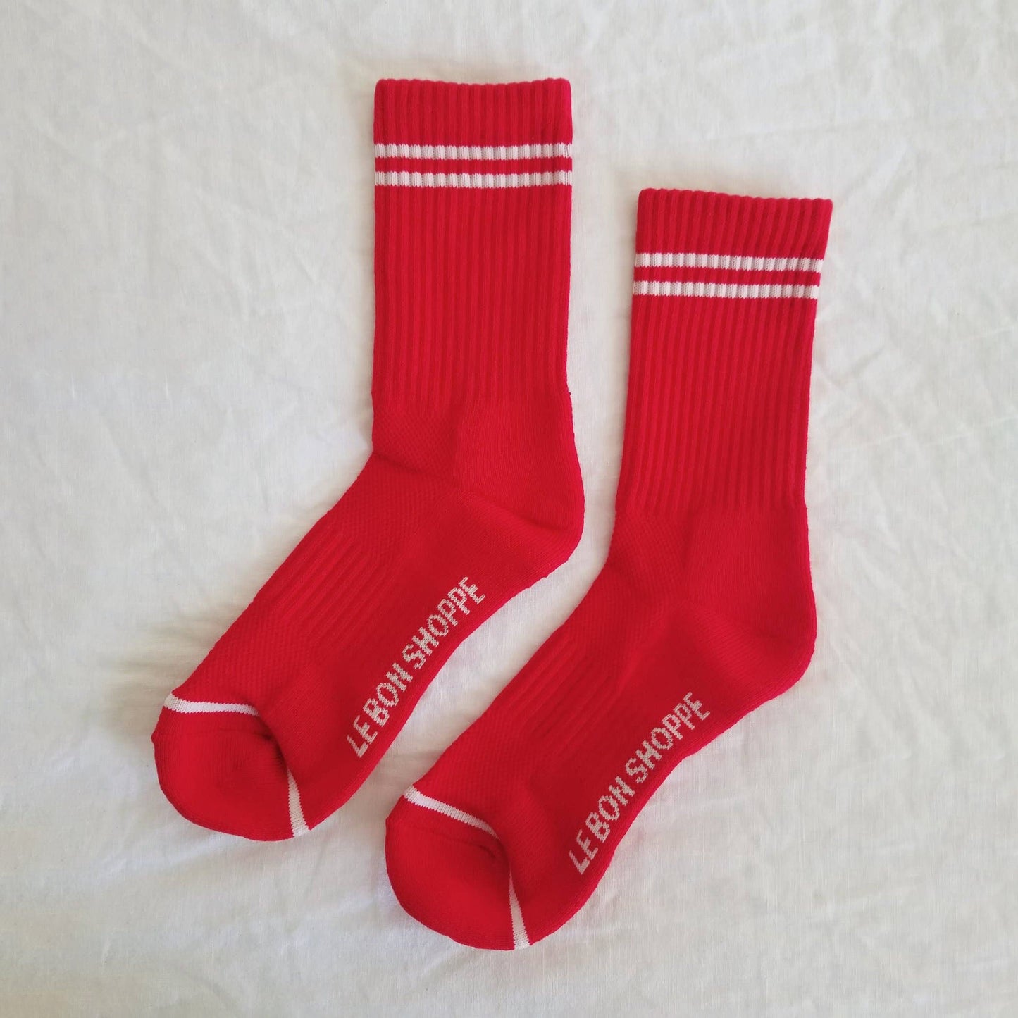 A pair of red ribbed crew socks with white stripes near the cuff, laid flat on a white fabric background, showing the Le Bon Shoppe branding on the sole and white toe detailing.
