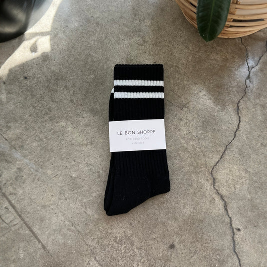 A pair of black extended boyfriend socks with white stripes, displayed on a textured surface, with a brand tag attached showing 'LE BON SHOPPE' and 'Christmas Sale'.