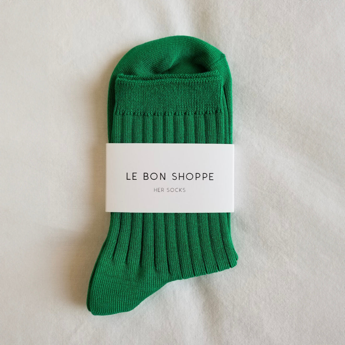 A pair of Kelly green ribbed crew socks neatly folded and wrapped with a white Le Bon Shoppe label, placed on a white fabric background.